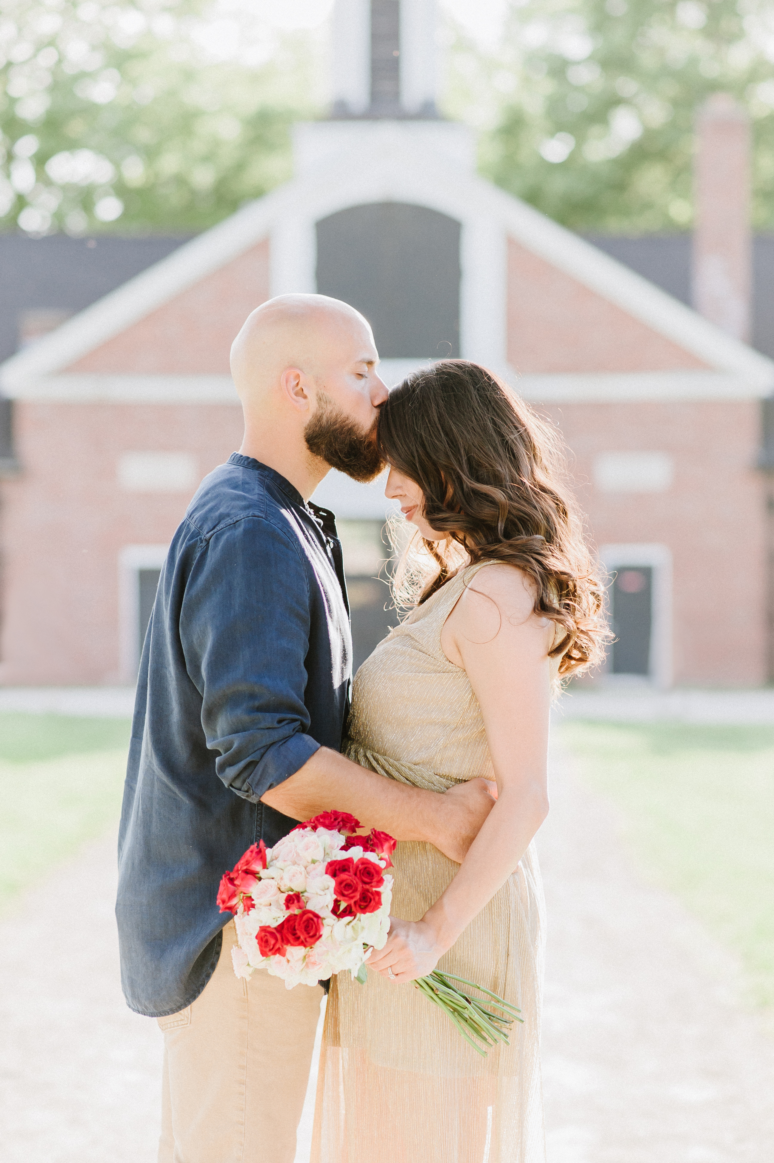 couple hugging in front of barn wearing gold and blue with red bouquet