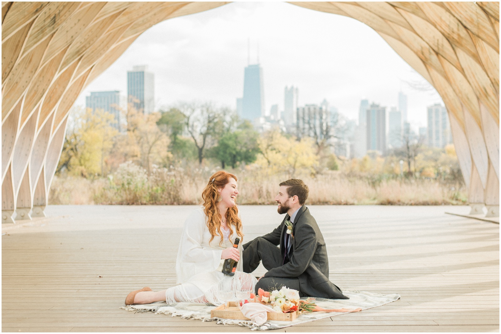 bride and groom sitting on picnic blanket drinking champagne