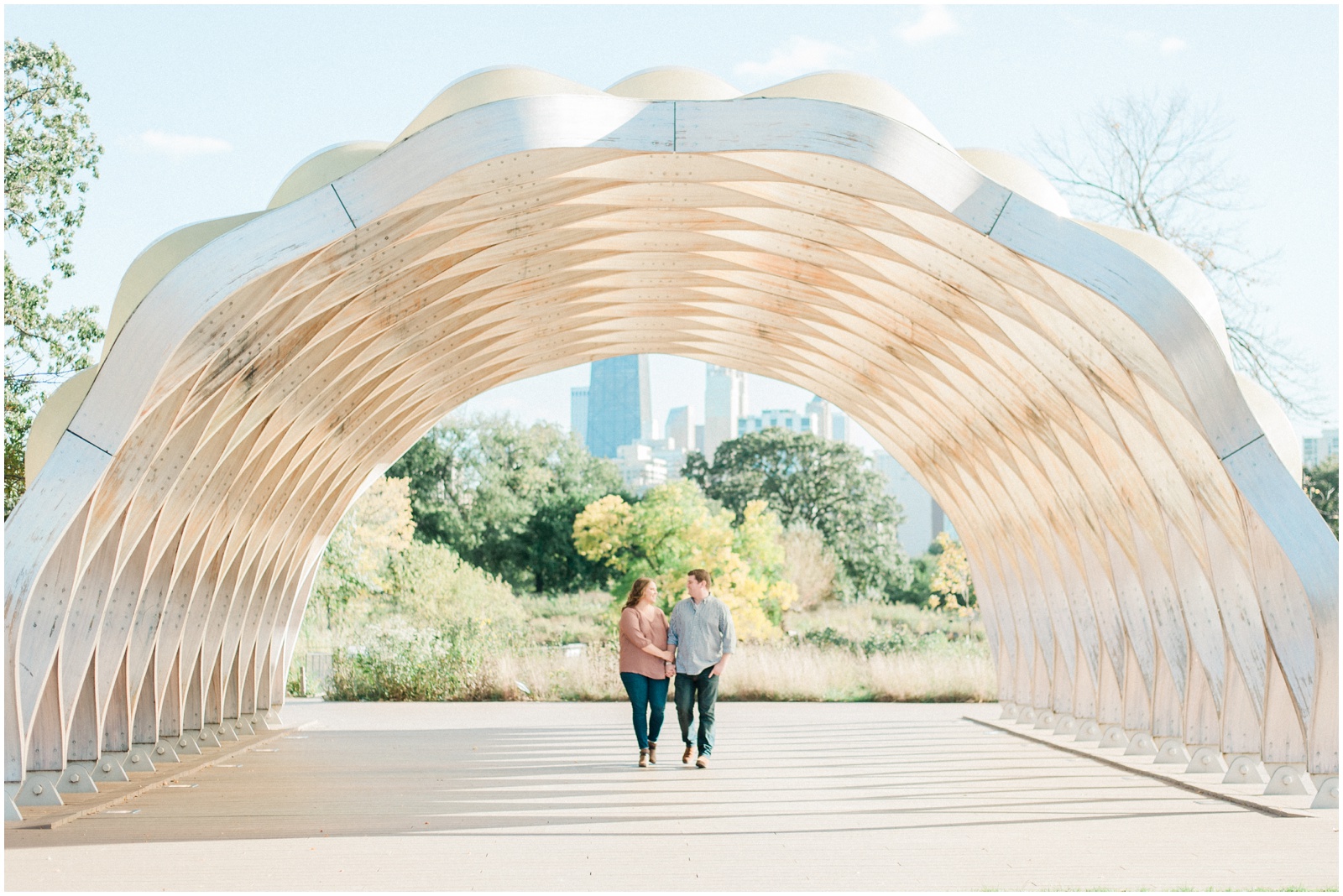 couple walking under architectural archway in park overlooking chicago skyline for engagement photos