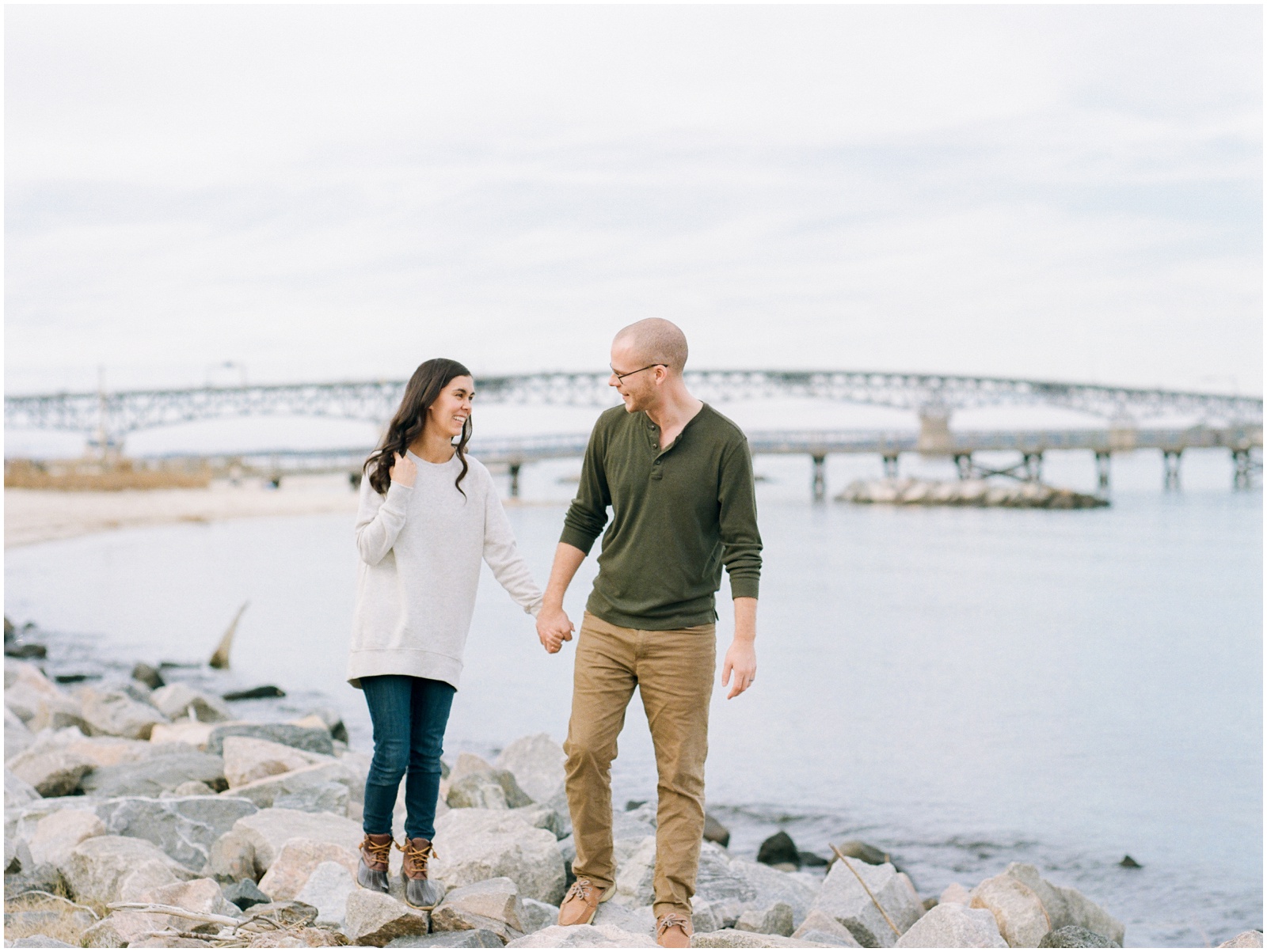 couple walking along rocks in front of a river and a bridge during engagement session